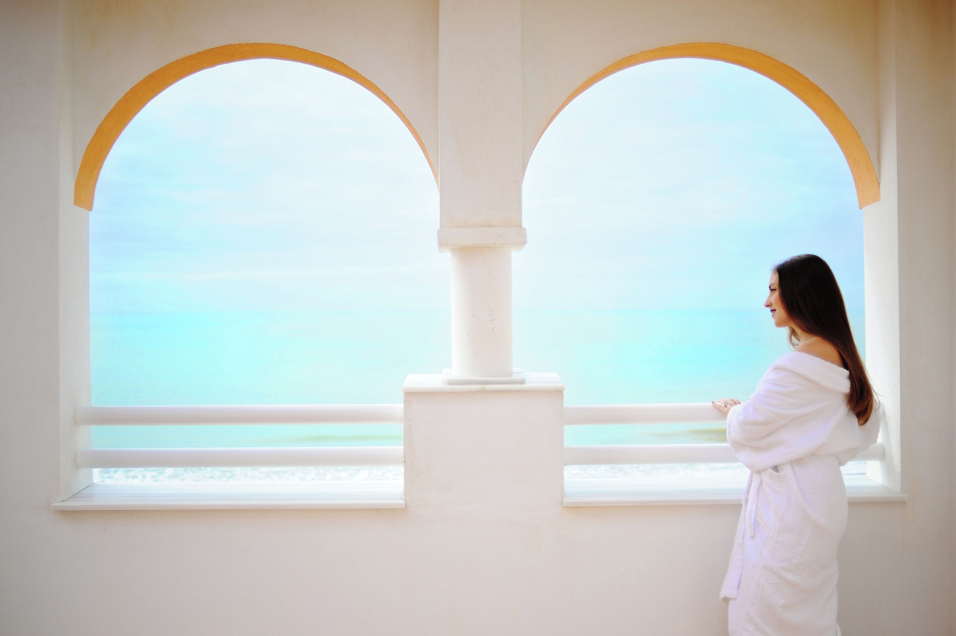 Una persona vestida de blanco de pie junto a una ventana con vista al mar.