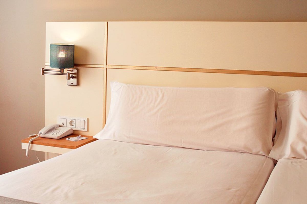 A hotel room with a bed, a bedside table with a phone, and a wall-mounted lamp.