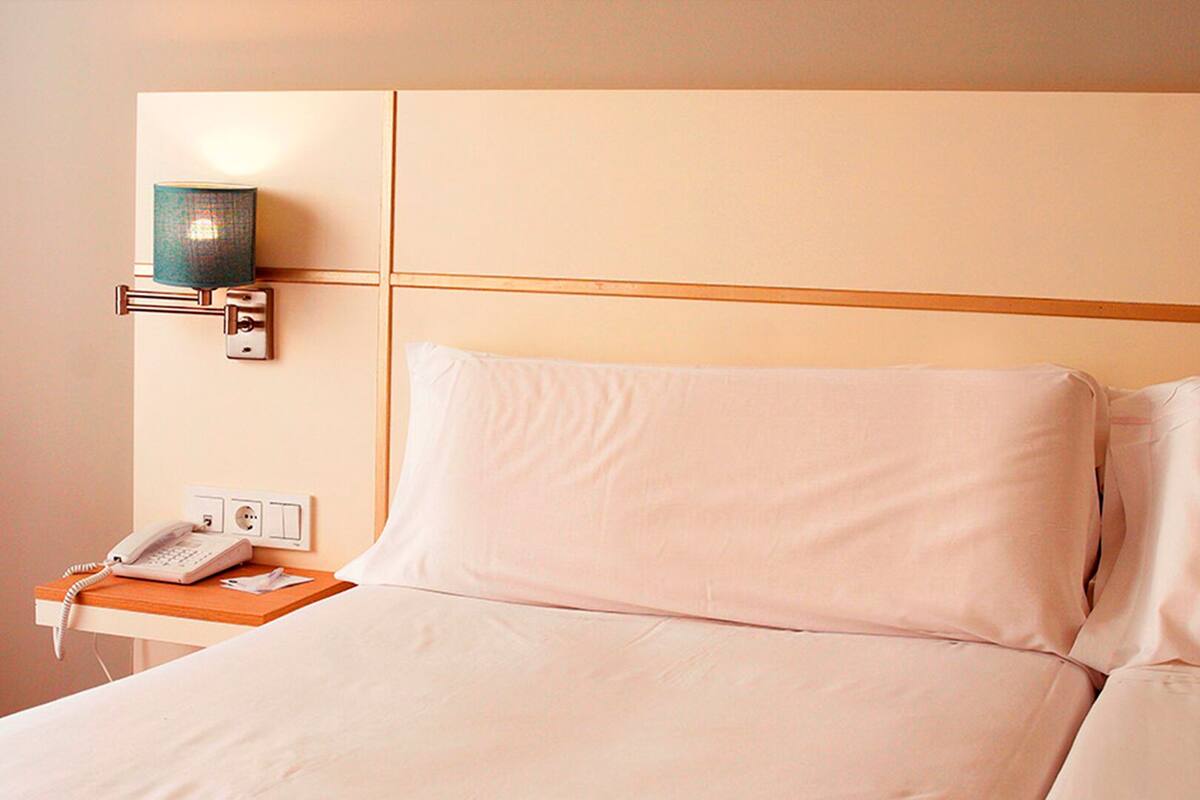 A hotel room with a bed, a bedside table with a telephone, and a wall-mounted lamp.