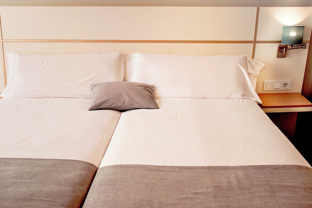 A double bed with two pillows, a bedside table with a lamp, and a wall-mounted light fixture.