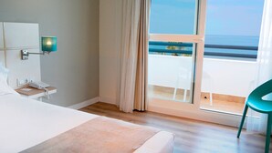 A hotel room with a bed, a bedside telephone, a nightstand, a window with curtains, and a view of the sea. - Hotel Vertice Chipiona Mar (Chipiona)