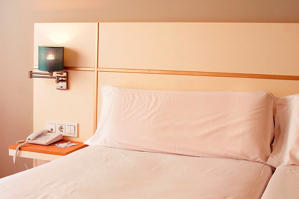 A hotel room with a bed, a bedside table with a telephone, and a wall-mounted lamp.
