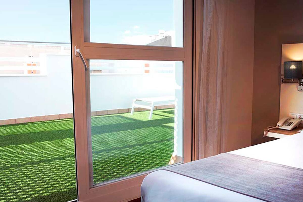 A hotel room with a large window, a bed, a bedside table with a phone, and a view of a green lawn and white chair outside.