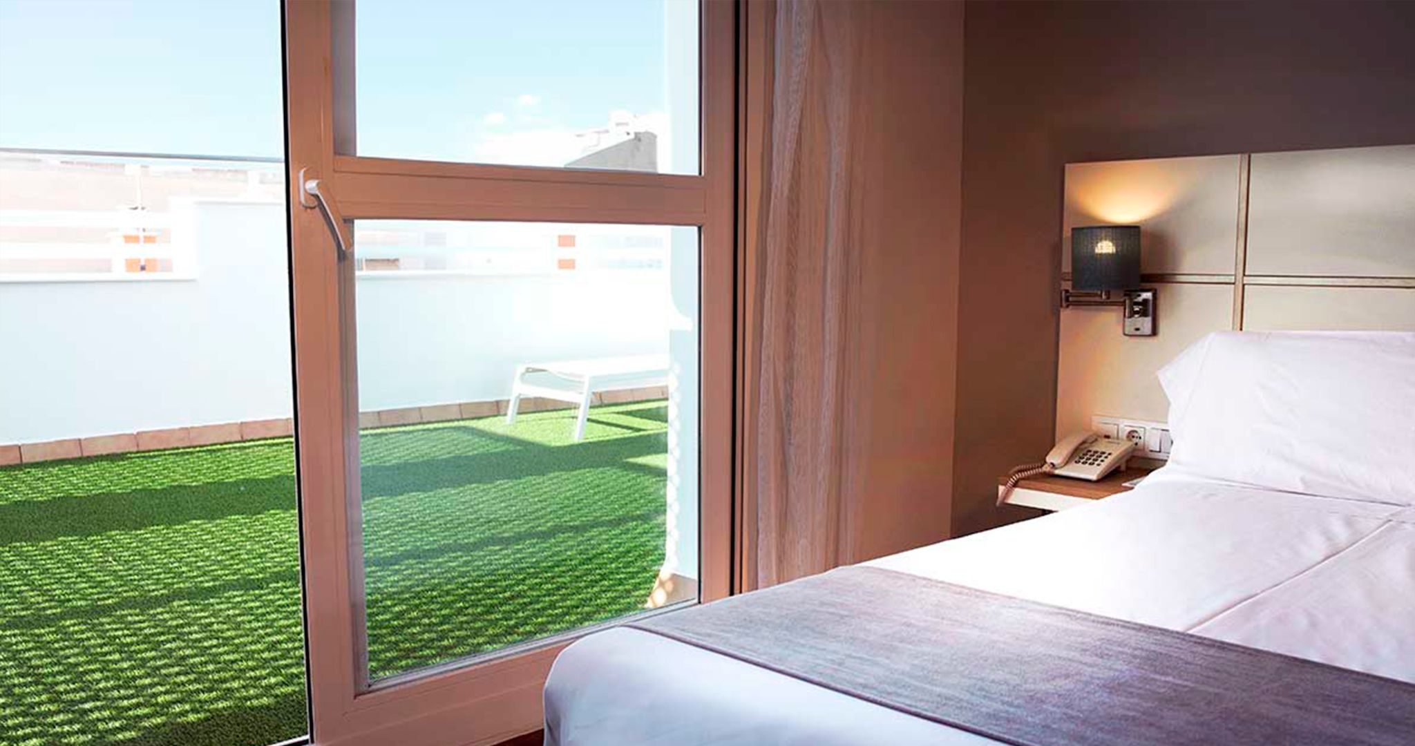 A hotel room with a large window, a bed, a bedside table with a phone, and a view of a green lawn and white chair outside.