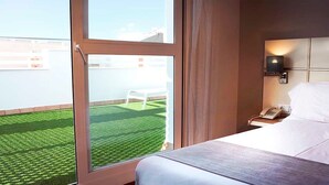 A hotel room with a large window, a bed, a bedside table with a phone, and a view of a green lawn and white chair outside. - Hotel Vertice Chipiona Mar (Chipiona)