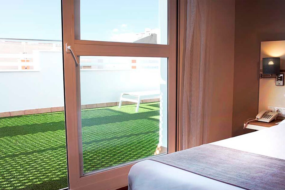 A hotel room with a large window, a bed, a bedside table with a phone, and a view of a green lawn and white chair outside.