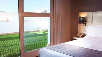 A hotel room with a large window, a bed, a bedside table with a phone, and a view of a green lawn and white chair outside.