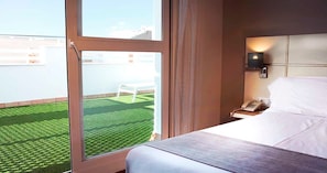 A hotel room with a large window, a bed, a bedside table with a phone, and a view of a green lawn and white chair outside. - Hotel Vertice Chipiona Mar (Chipiona)