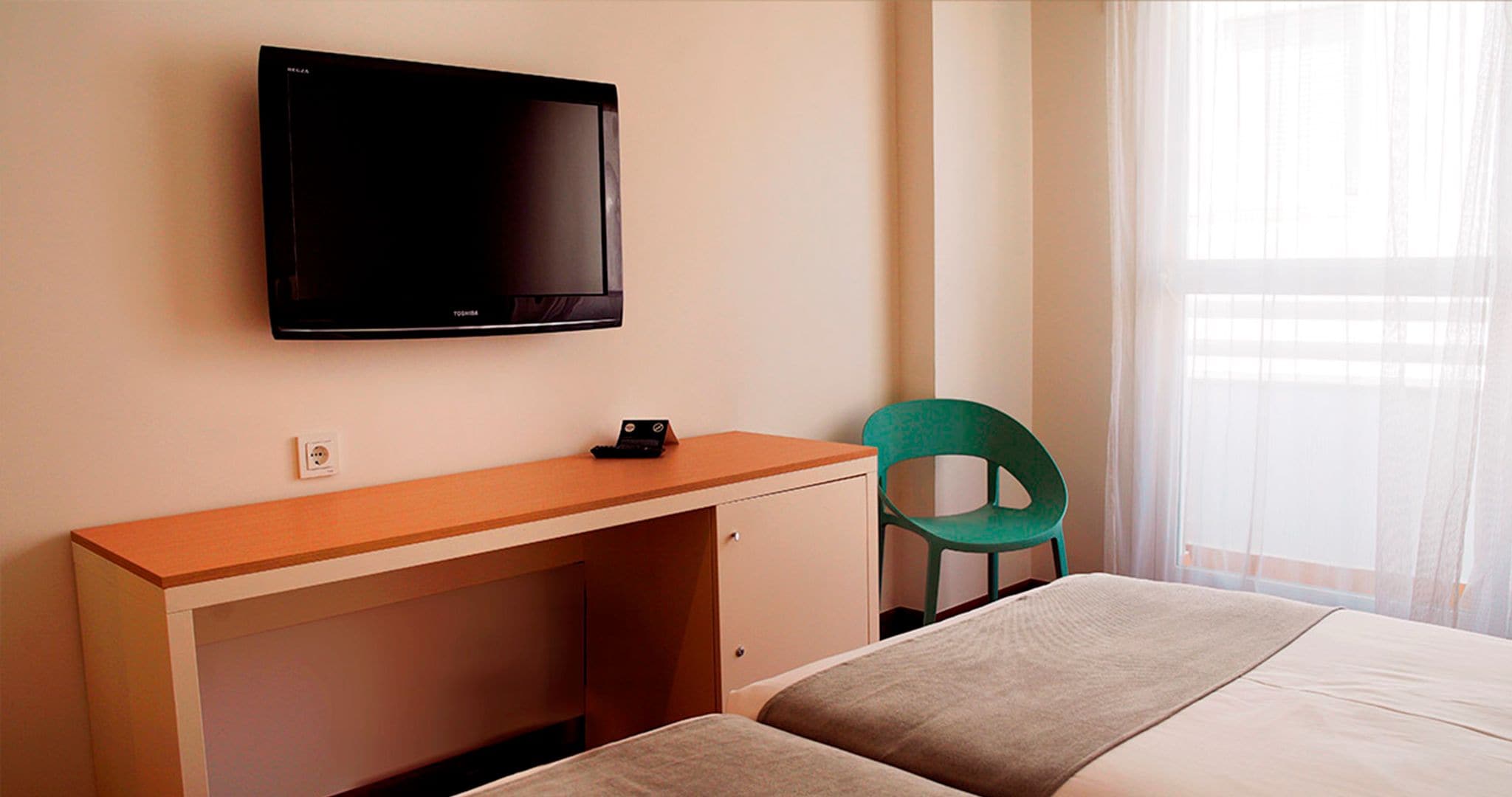 Habitación de hotel con una cama, un escritorio con computadora, una silla verde y un televisor montado en la pared.