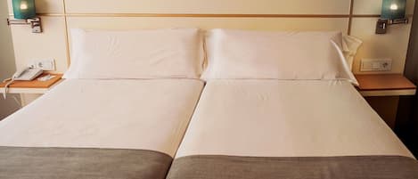 Un lit double avec du linge de lit blanc, deux tables de chevet, un téléphone et des lampes fixées au mur.