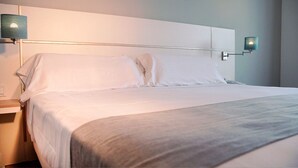 Cama bem arrumada com lençóis brancos e manta cinza, mesa de cabeceira com abajur, e luminária fixada na parede.