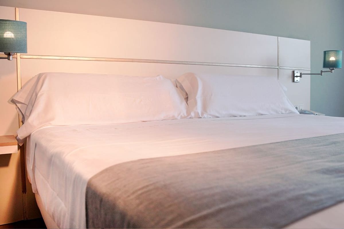 A neatly made bed with white linens and a gray throw blanket, a bedside table with a lamp, and a wall-mounted light fixture.