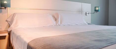 A neatly made bed with white linens and a gray throw blanket, a bedside table with a lamp, and a wall-mounted light fixture.