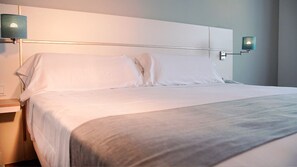 A neatly made bed with white linens and a gray throw blanket, a bedside table with a lamp, and a wall-mounted light fixture. - Hotel Vertice Chipiona Mar (Chipiona)