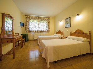 A room with two single beds, a wooden desk with a chair, a radiator, a television on the wall, and a window with floral curtains. - El Mirador (Puertolas)