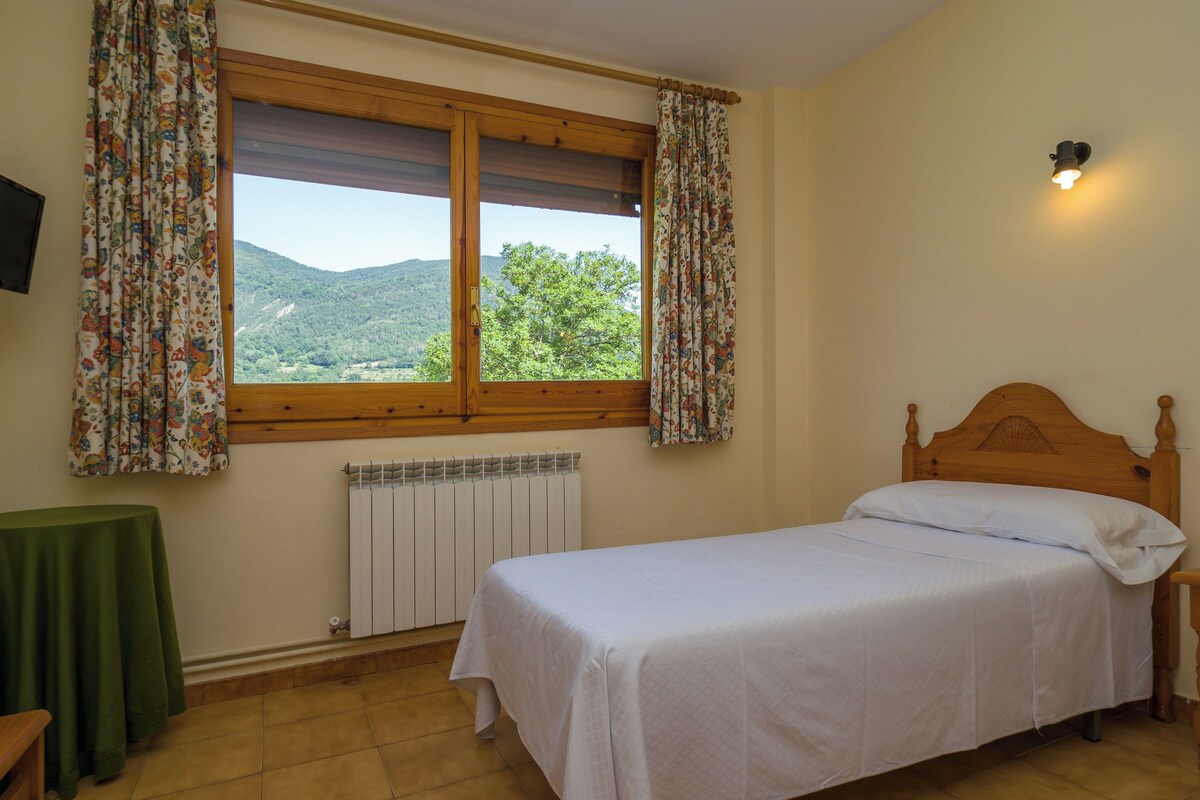 Quarto com uma cama de solteiro, janela com cortinas, radiador e uma mesinha.