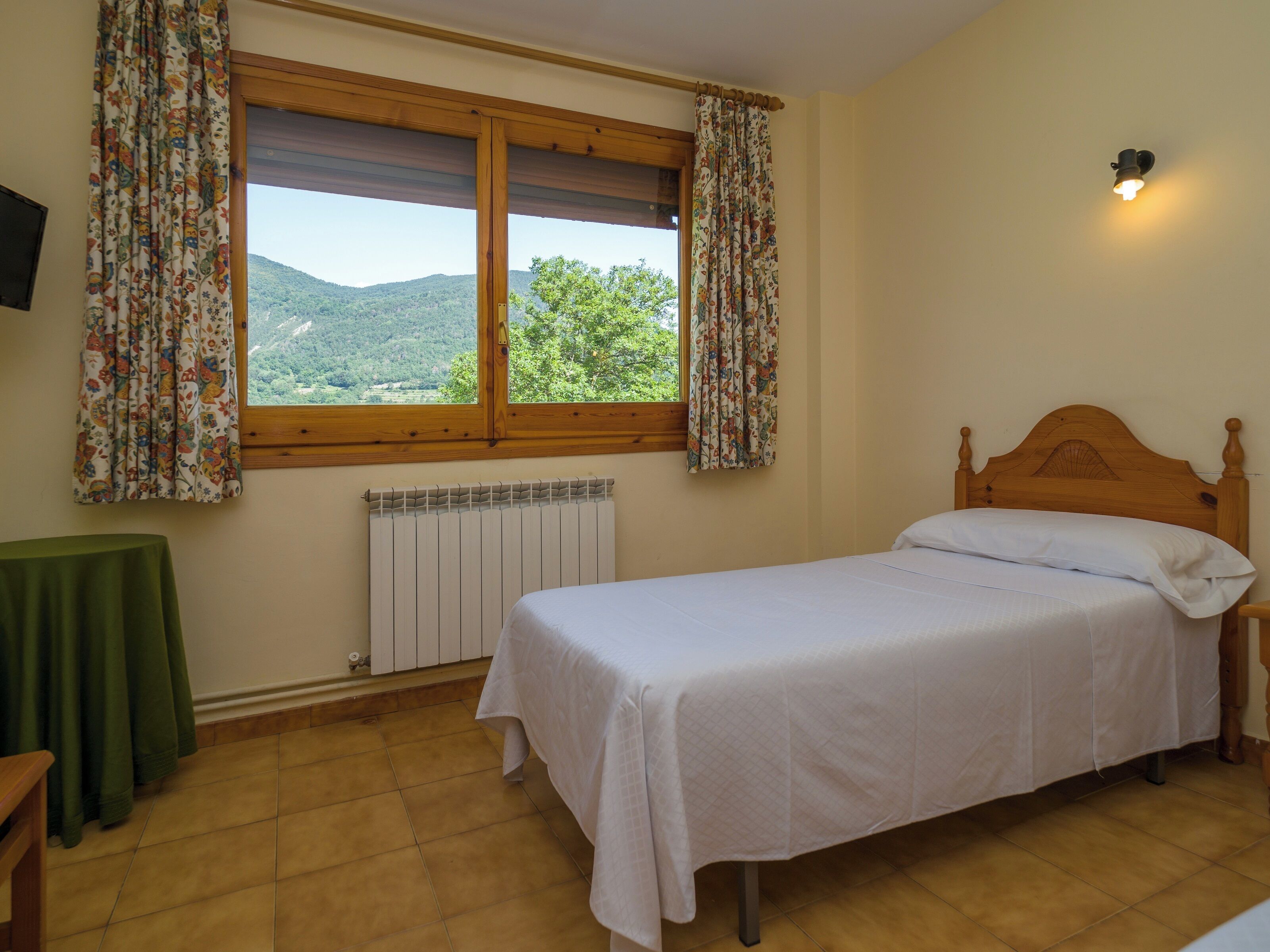 Ein Schlafzimmer mit einem Einzelbett, einem Fenster mit Vorhängen, einem Heizkörper und einem kleinen Tisch.