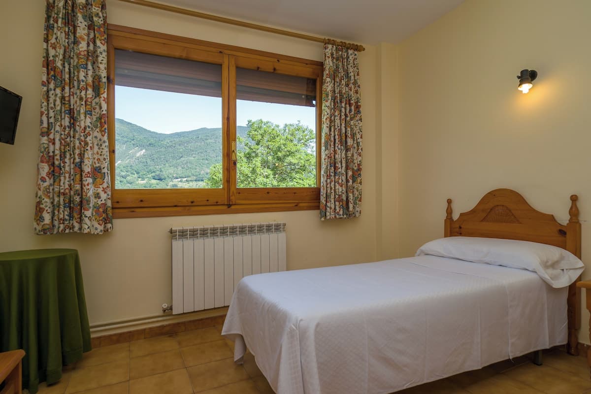 A bedroom with a single bed, a wooden headboard, a window with floral curtains, a radiator, and a small table with a green tablecloth.