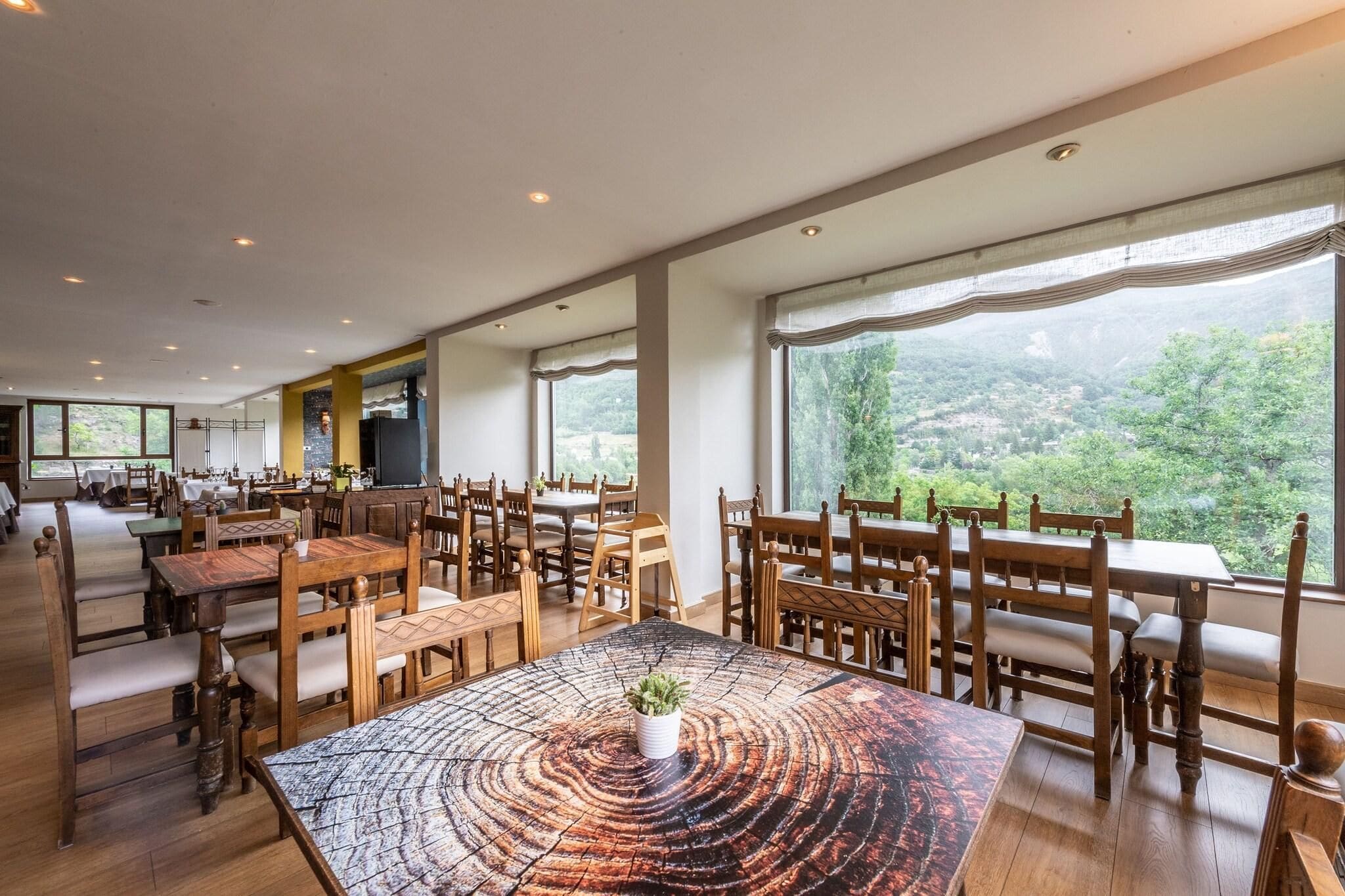 A spacious dining area with wooden tables and chairs, large windows offering a view of a green landscape.