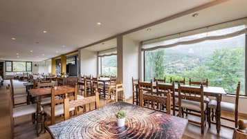 A spacious dining area with wooden tables and chairs, large windows offering a view of a green landscape.