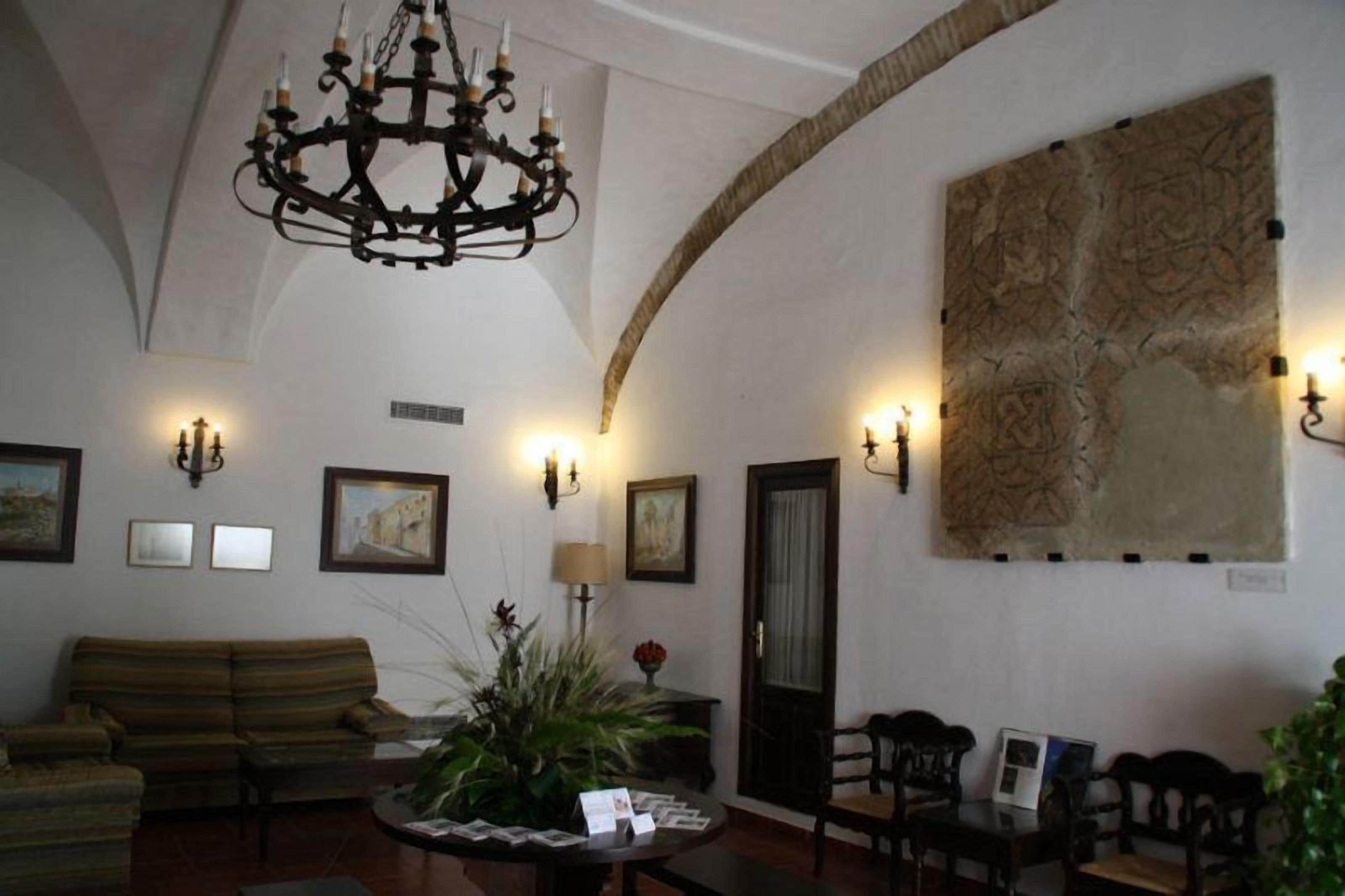 A spacious lobby with a high arched ceiling, a chandelier, wall-mounted sconces, a large abstract painting, a sofa, a coffee table with magazines, and a door.