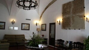 A spacious lobby with a high arched ceiling, a chandelier, wall-mounted sconces, a large abstract painting, a sofa, a coffee table with magazines, and a door. - Convento de San Francisco (Vejer de la Frontera)