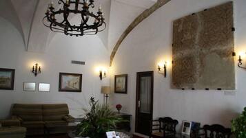A spacious lobby with a high arched ceiling, a chandelier, wall-mounted sconces, a large abstract painting, a sofa, a coffee table with magazines, and a door.