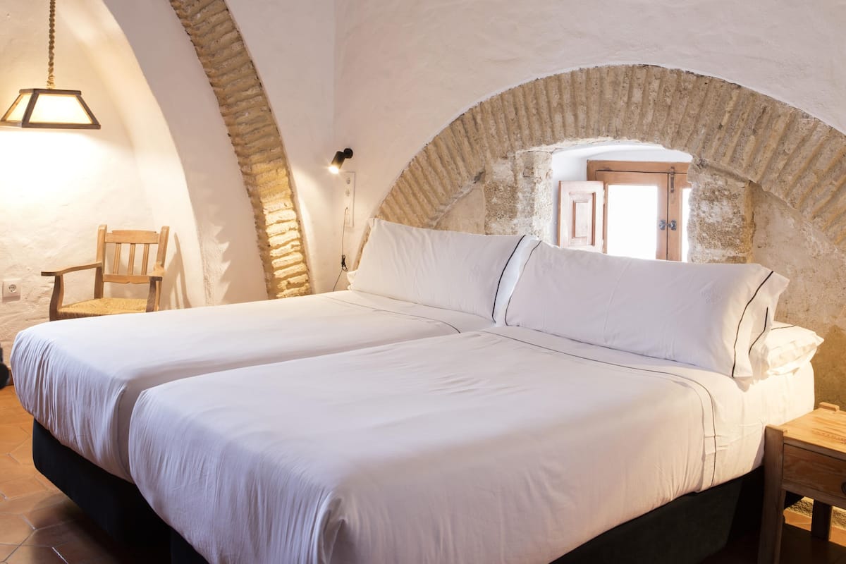 Ein Schlafzimmer mit zwei Betten, einem Holzstuhl, einer Pendelleuchte und einem Steinbogen.