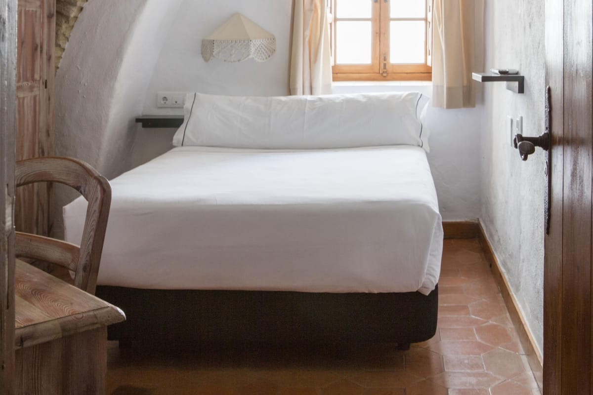Ein Schlafzimmer mit einem Bett, einem fensterbelaubten Fenster, einer Holztür und einem Fliesenboden.