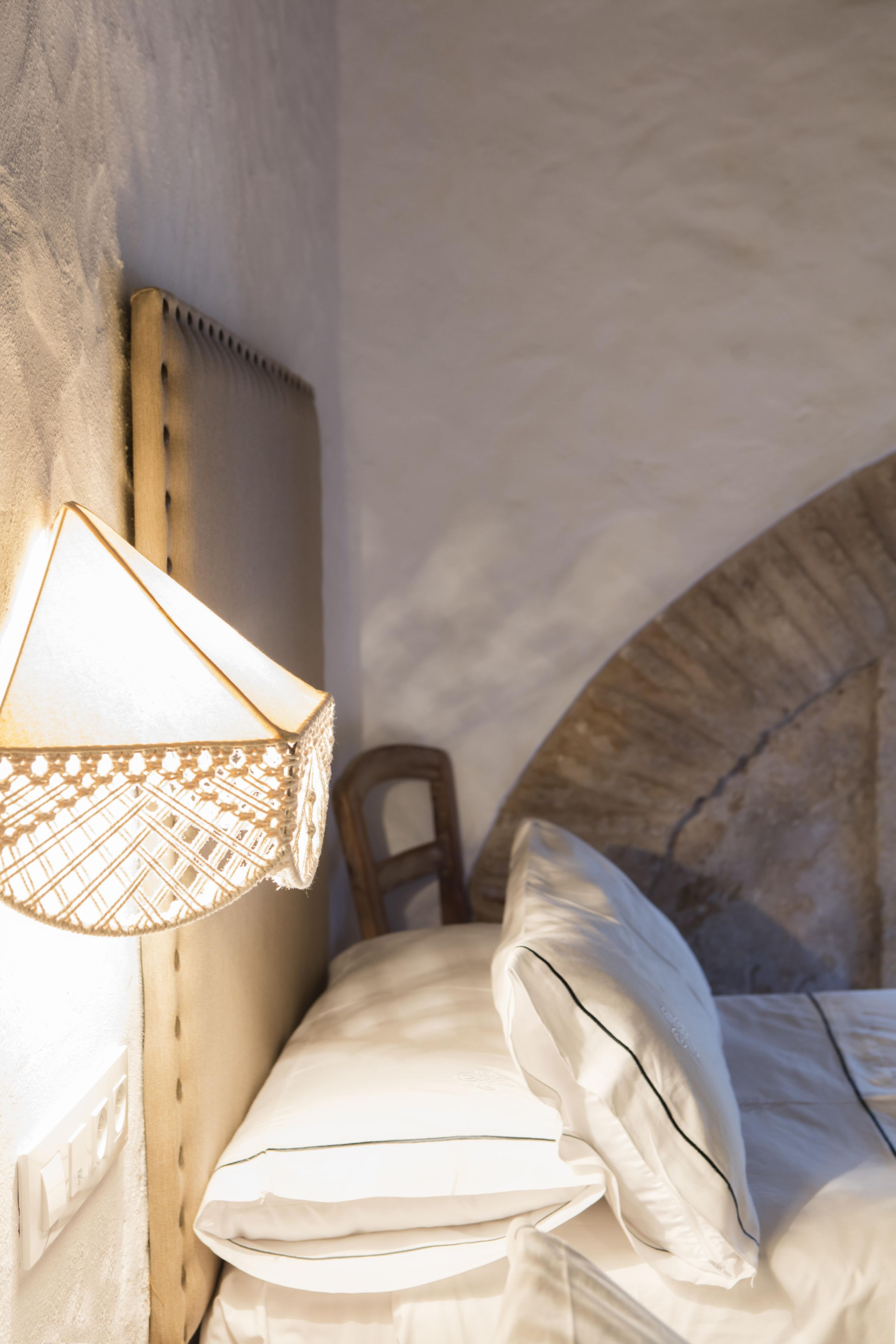 A bedroom with a headboard, a bedside lamp, a bed with pillows, and a chair.