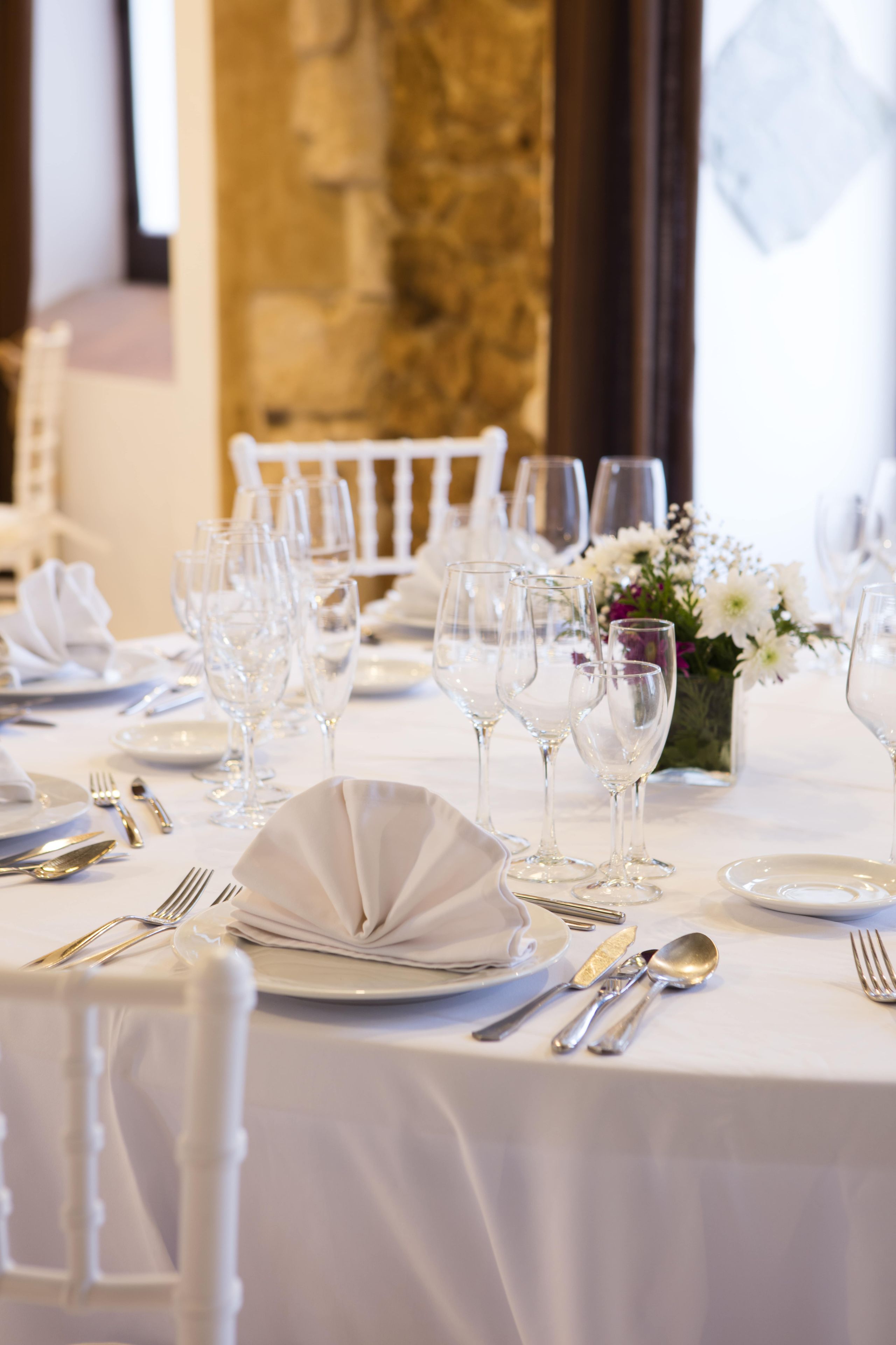 Mesa de comedor formal cubierta con mantel blanco, cubiertos y copas de vino, con un jarrón de flores en el centro.
