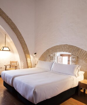 A bedroom with two beds, a wooden chair, a hanging light fixture, and a stone arch. - Convento de San Francisco (Vejer de la Frontera)