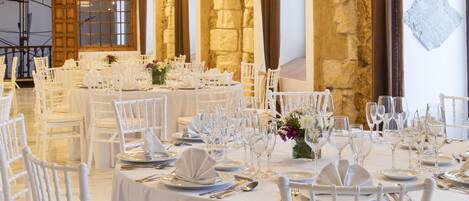 Un salón de banquetes con mesas y sillas blancas, paredes de piedra y lámparas colgantes.