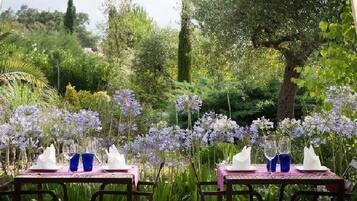 Un área de comedor al aire libre con mesas dispuestas para dos, rodeada de exuberante follaje y flores moradas.