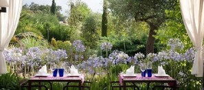 An outdoor dining area with tables set for two, surrounded by lush greenery and purple flowers. - Aatu (Forallac)