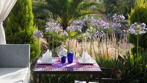 An outdoor dining table set with white napkins, blue glassware, and a patterned tablecloth, surrounded by lush greenery and purple flowers. - Aatu (Forallac)