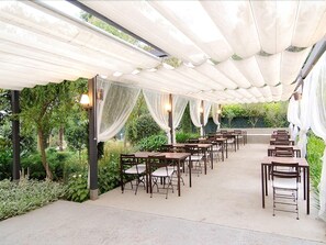 An outdoor dining area with tables and chairs under a white canopy, surrounded by greenery. - Aatu (Forallac)