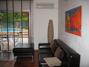 Un salón con un sofá de cuero negro, un cuadro abstracto de gran tamaño y vistas a una piscina a través de una ventana con persianas.