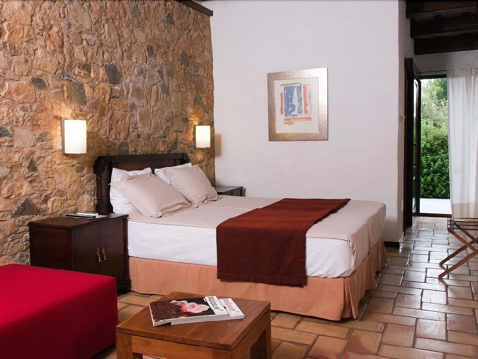 A bedroom with a stone wall, a bed with a red blanket, a wooden nightstand, a red chair, and a wooden table with a magazine.
