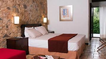 A bedroom with a stone wall, a bed with a red blanket, a wooden nightstand, a red chair, and a wooden table with a magazine.