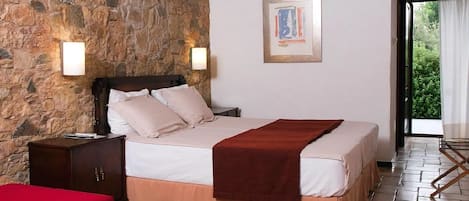 Un dormitorio con pared de piedra, una cama con manta roja, una mesita de noche de madera, una silla roja y una mesa de madera con una revista.