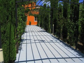 Un camino conduce a un edificio de fachada naranja, rodeado de follaje y una estructura de pérgola metálica.