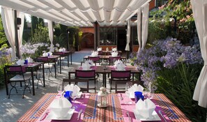 Un espace de restauration en plein air, avec des tables disposées pour deux personnes, recouvert d’un auvent blanc et entouré d’une végétation luxuriante et de fleurs violettes.