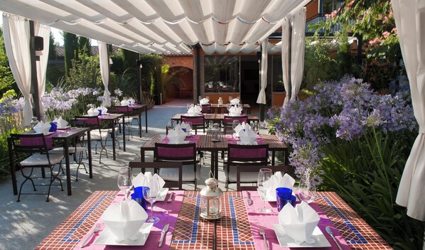 An outdoor dining area with tables set for two, covered by a white canopy, surrounded by lush greenery and purple flowers. - Aatu (Forallac)
