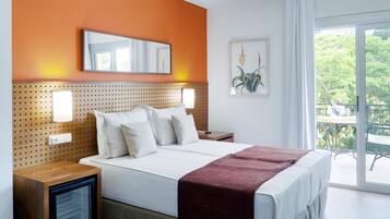 Habitación de hotel con cama, mesita de noche, pequeño refrigerador y una ventana grande con vistas a árboles.