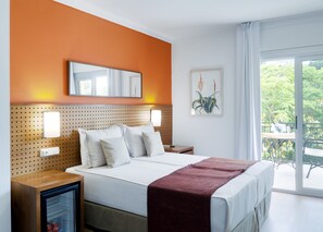 A hotel room with a bed, a bedside table, a nightstand, a small refrigerator, and a large window with a view of trees. - Aatu (Forallac)