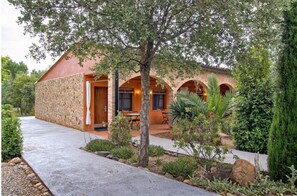 A stone building with a covered patio, arched windows, and a garden with various plants and a tree. - Aatu (Forallac)