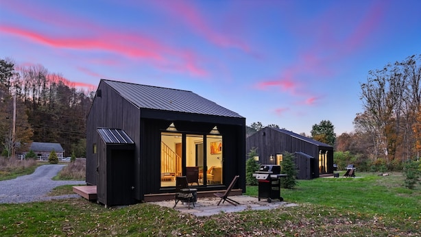Una cabaña moderna de color oscuro, con un gran ventanal y ventanas, un patio con mobiliario exterior y una zona de barbacoa.