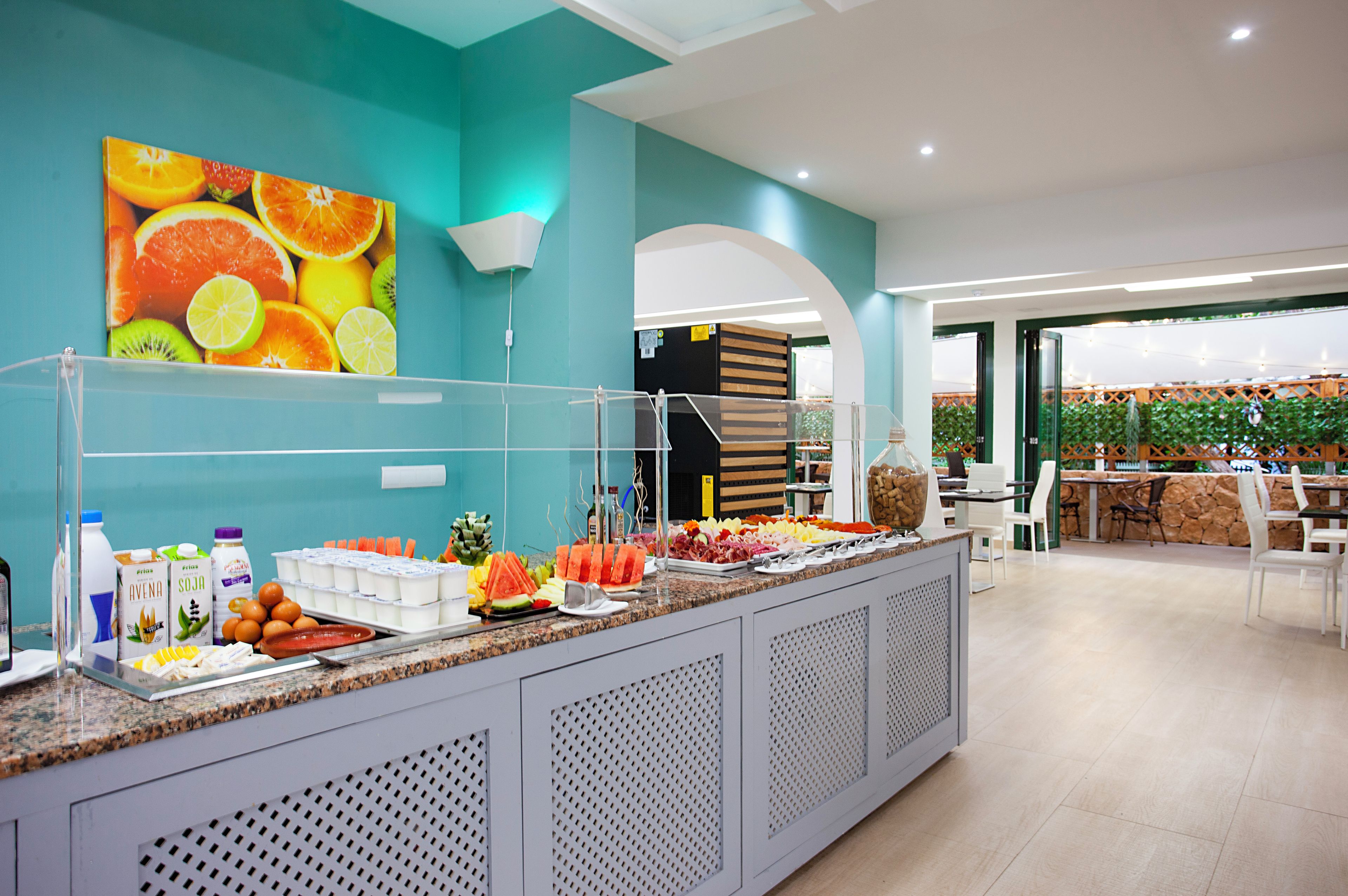 A buffet area with a variety of fruits and beverages, a large fruit painting on the wall, and a seating area with tables and chairs.