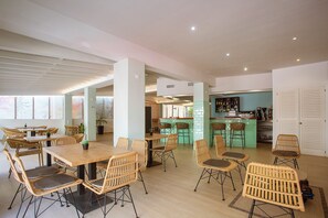 A spacious dining area with high tables and wicker chairs, a bar area with stools, and a view of the city through large windows. - Hotel Rosales Formentera (Formentera)
